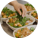 A hand adding fresh cilantro to a plant-based meal bowl filled with crispy tofu, greens, and vegetables, served with a side of dipping sauce in an eco-friendly container.