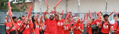 A large group of diverse young people wearing matching red shirts and wristbands enthusiastically raising red inflatable cheering sticks outdoors, appearing to celebrate or support an event.