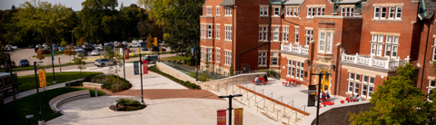 The University of Guelph Lang building with outdoor patio seating areas where students are sitting at red tables. The scene shows accessible pathways with ramps and handrails, landscaped greenery, and a parking lot in the background under a clear sky.