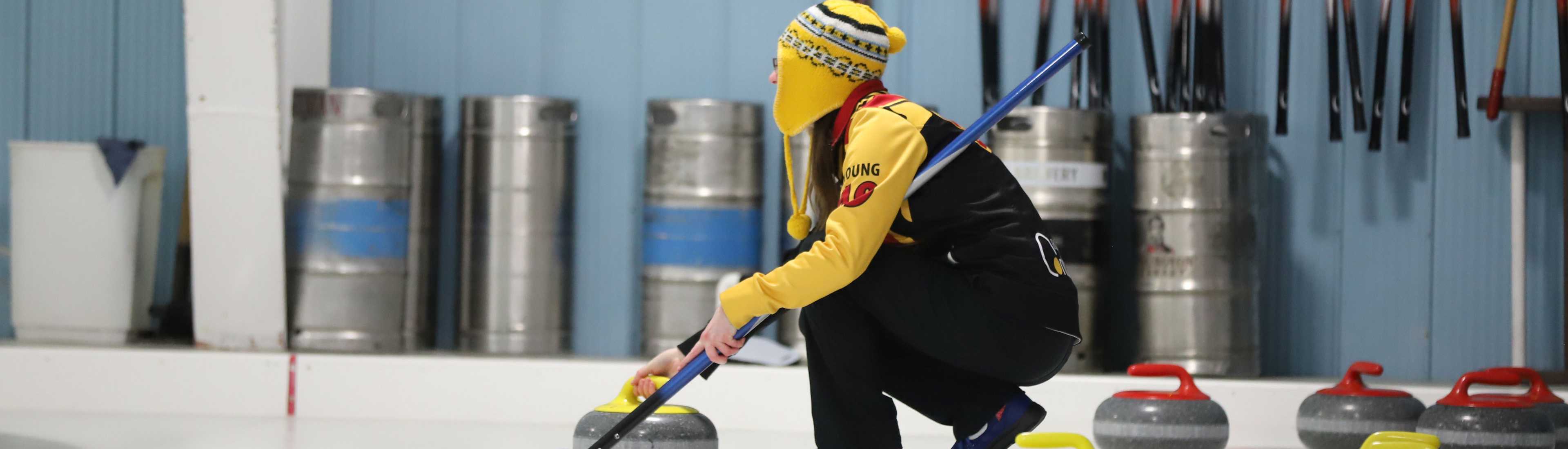 Curler in yellow and black gear preparing to deliver a curling stone on the ice at the EAA AGM & Bonspiel event, with equipment and curling stones in the background.