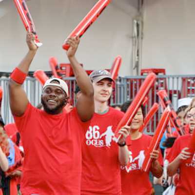 A large group of diverse young people wearing matching red shirts and wristbands enthusiastically raising red inflatable cheering sticks outdoors, appearing to celebrate or support an event.