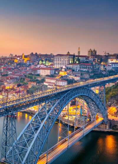 A stunning view of the Luís I Bridge spanning the Douro River in Porto, Portugal, at sunset, with the city's historic architecture beautifully illuminated in the background.