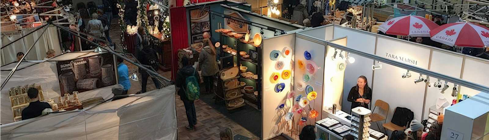 Booths at the Fair November Craft Show in the University Centre, Guelph, showcasing handmade Canadian goods. Artisans display pottery, colorful glass art, wooden items, and jewelry, while shoppers explore the stalls under Canadian flag umbrellas.