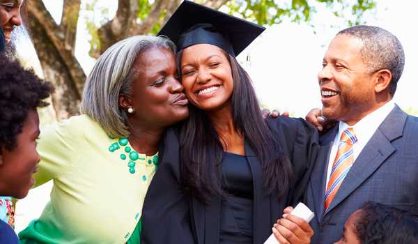 Smiling graduate in cap and gown embraces her proud parents as they celebrate together, with the mother kissing her daughter on the cheek.