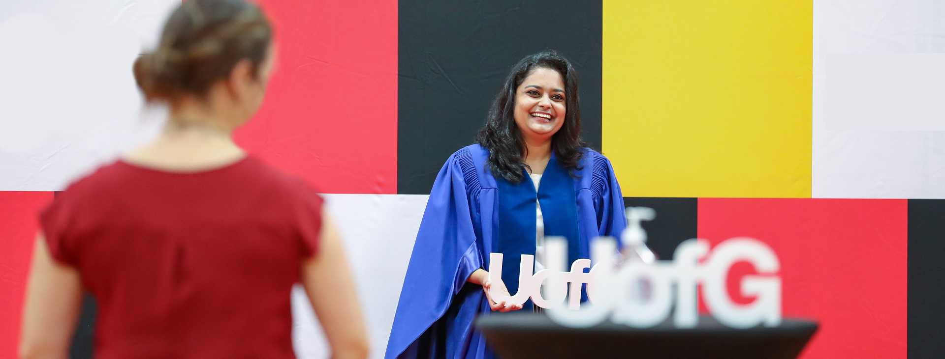 A smiling woman in a blue graduation gown stands in front of a colorful background with red, black, yellow, and white blocks. She is holding a white cutout of the letters 'U of G'. Another person, seen from behind and wearing a red top, is facing her.