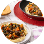 A plate of rich beef stew with tender carrots, pearl onions, and herbs served over creamy mashed potatoes, with a skillet of the dish in the background on a red placemat.