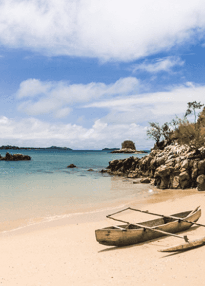 Wooden outrigger canoe resting on a sandy beach with turquoise waters, rocky shoreline, and a partly cloudy sky on the coast of Madagascar.
