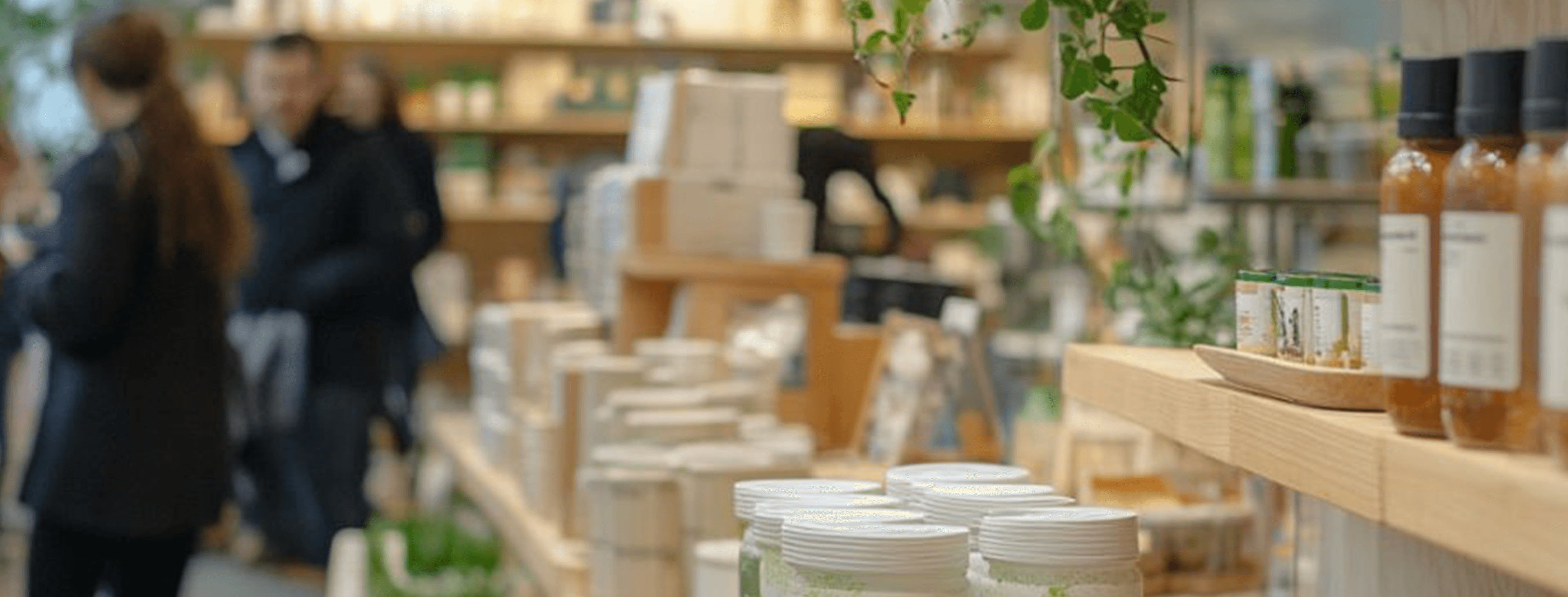 A well-lit store with wooden shelves displaying neatly arranged products, including bottles, jars, and boxes. The foreground features a row of amber-colored bottles with black caps and white labels. In the background, blurred shoppers browse the store, which is decorated with green plants and natural materials, creating a warm and inviting atmosphere.