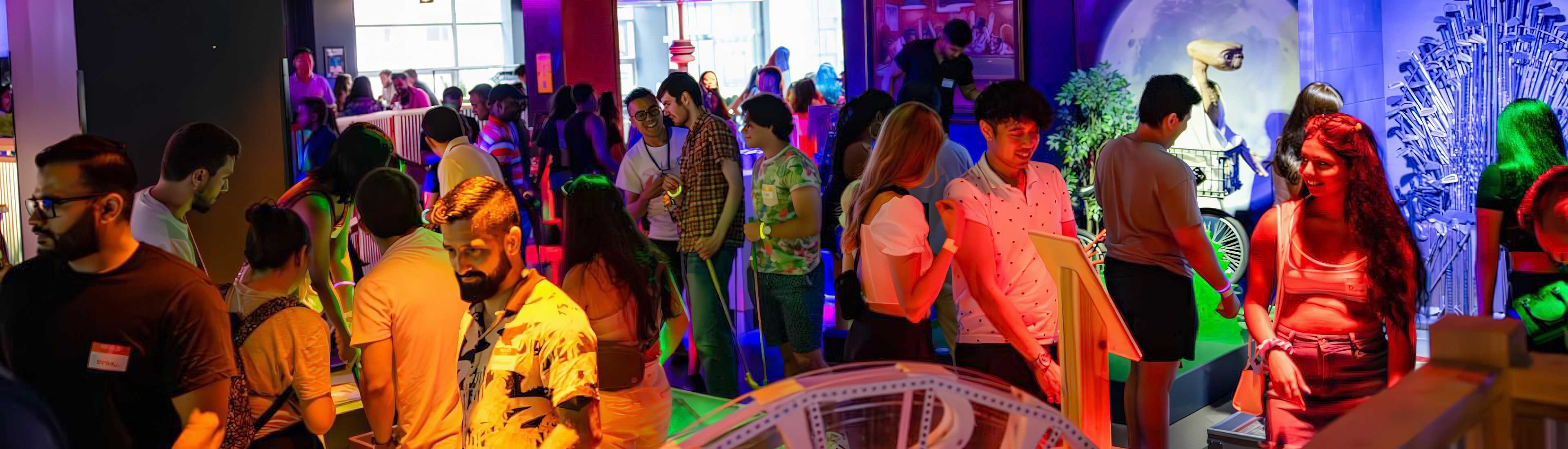 A lively crowd at an indoor mini-golf event with vibrant lighting. Participants are seen enjoying the game, interacting in groups, and exploring the colorful setting.