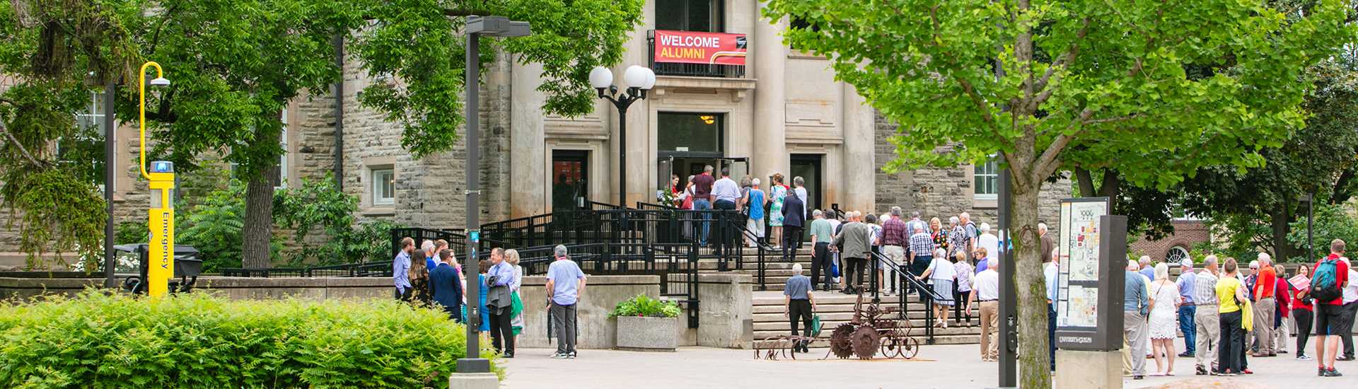 A group of people gathering outside a building with a "Welcome Alumni" banner at the University of Guelph. The scene includes a bright yellow emergency phone, lush green trees, and a map of the campus.