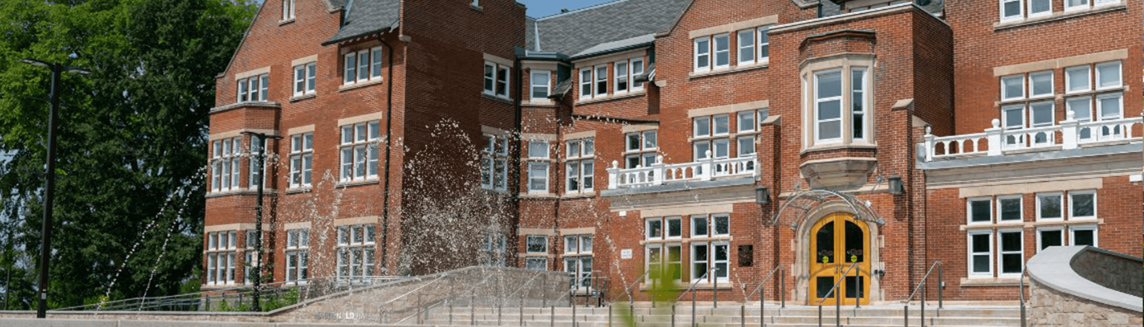 Macdonald Hall at the University of Guelph, a historic red-brick building with large windows and a decorative fountain in the foreground, surrounded by greenery.