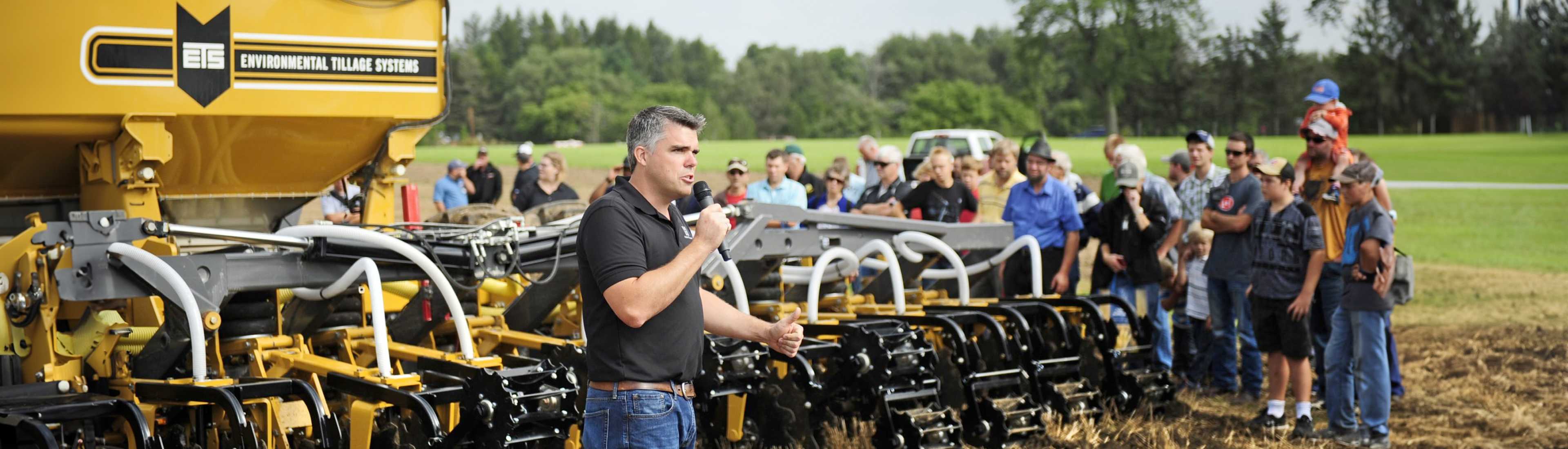 A presenter speaks into a microphone at the Outdoor Canada Farm Show, demonstrating agricultural equipment to a crowd of attendees.