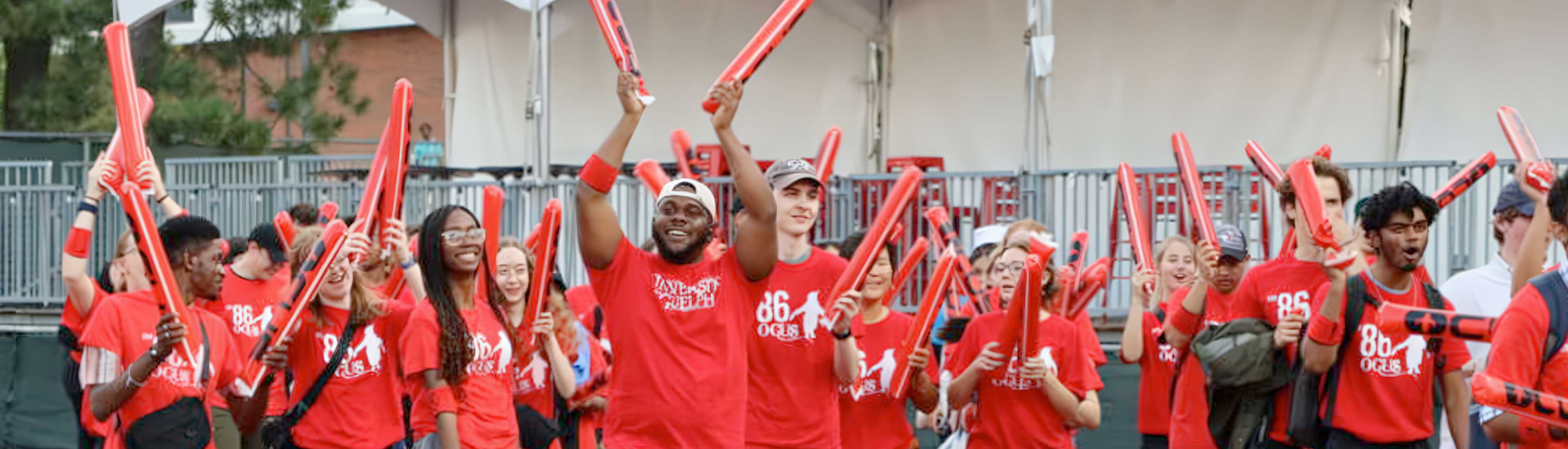 Group of university of Guelph students wearing red shirts and cheering during an outdoor orientation event, holding inflatable red sticks and smiling.