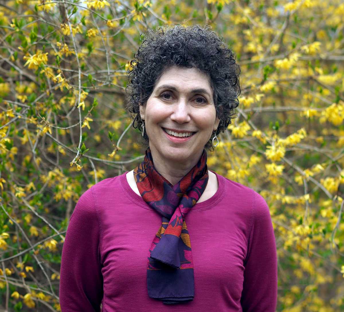 Barbara Salsberg Mathews, smiling and wearing a magenta top with a colorful scarf, standing outdoors in front of a background filled with yellow flowering branches.