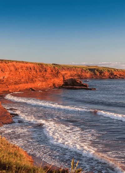 Scenic view of red sandstone cliffs along the coastline of Prince Edward Island, Canada, with ocean waves crashing against the shore under a clear blue sky.