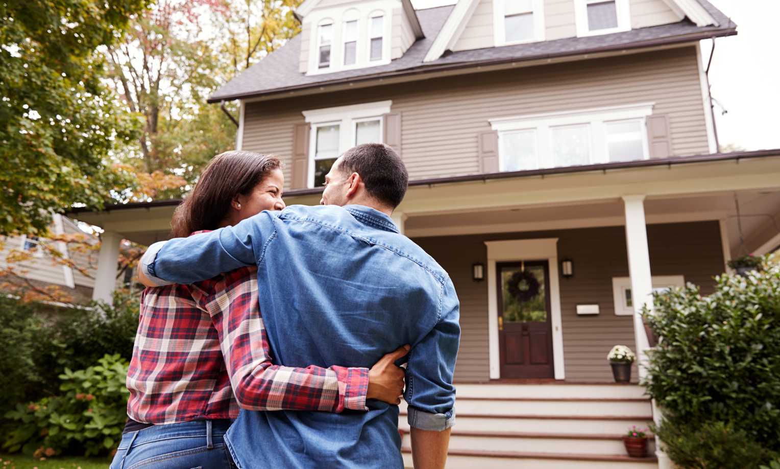 A couple embracing with their backs to the camera, looking at their new home on a sunny day.