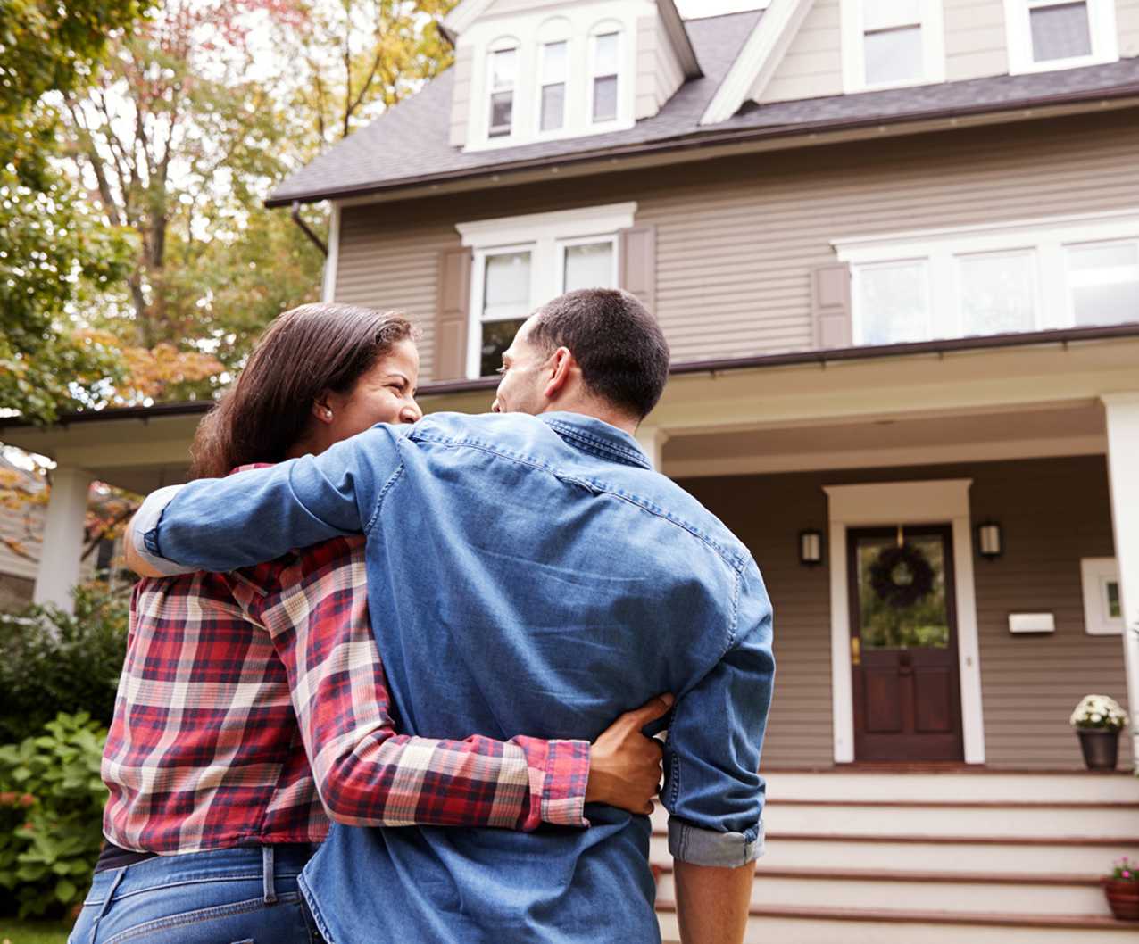 A couple stands arm in arm, smiling and looking at their new home, a two-story house with a front porch and steps, surrounded by trees and greenery.