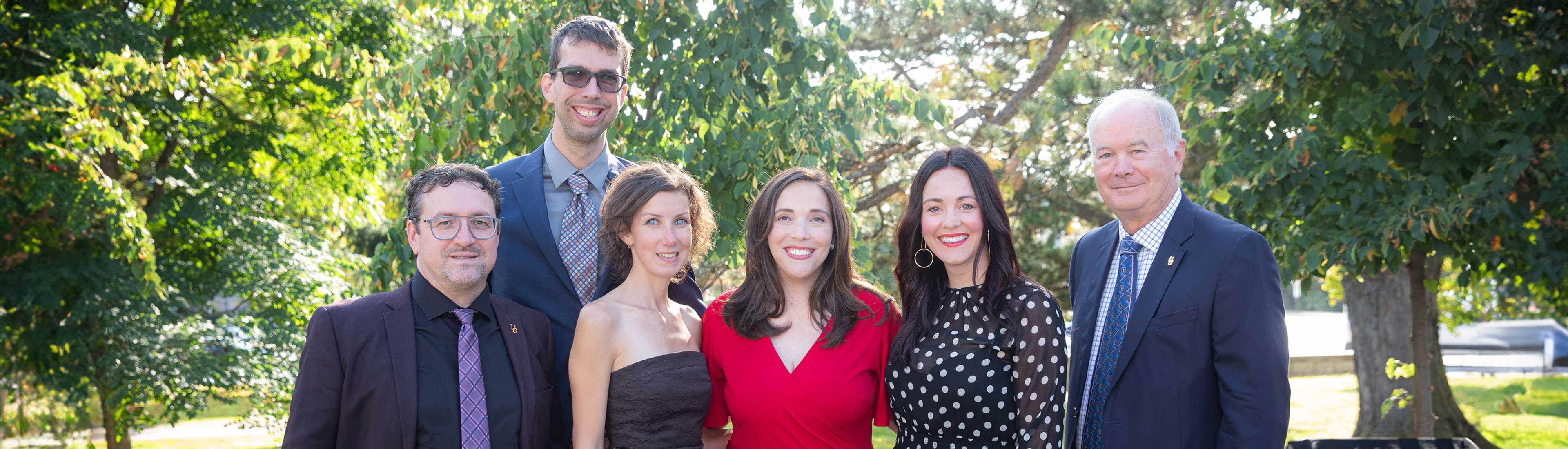 Group photo of University of Guelph staff at an outdoor event, featuring six individuals dressed in formal attire, standing in front of a lush green background.