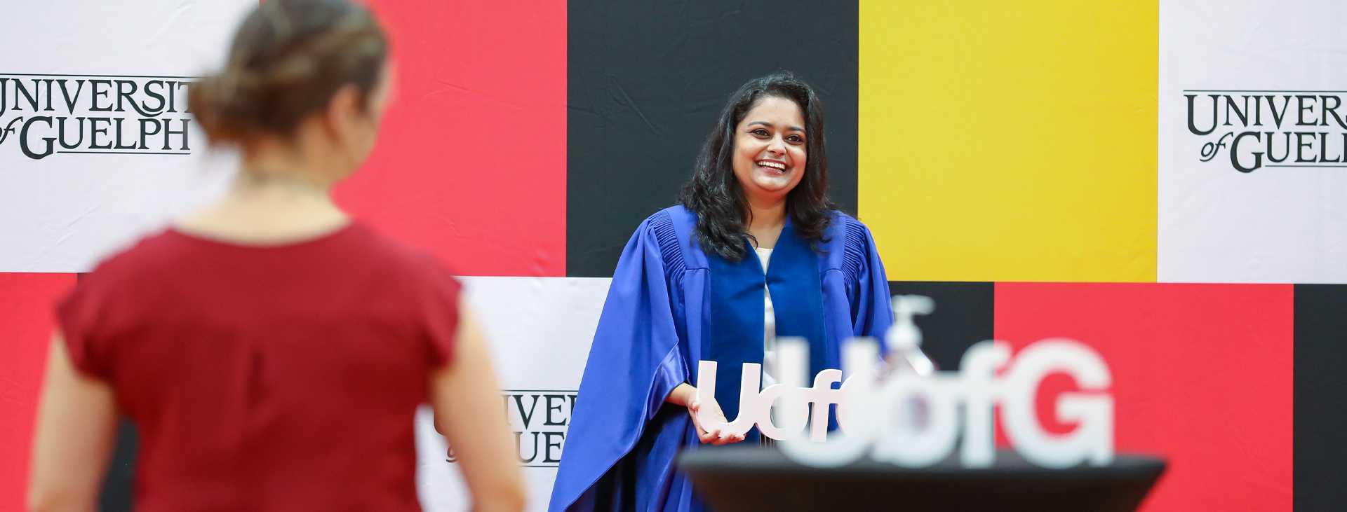 Smiling graduate in a blue gown posing for a photo at a University of Guelph graduation event, with a colorful backdrop and a 'U of G' sign in the foreground.