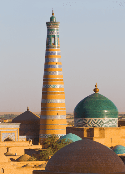 A scenic view of the Islam Khoja Minaret and surrounding domes in Khiva, Uzbekistan, bathed in warm sunlight. The tall, slender minaret is adorned with intricate blue, white, and orange tile patterns, while the nearby domes feature vibrant turquoise hues, reflecting the region's rich Islamic architecture.