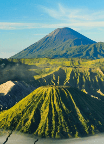 Scenic view of Mount Bromo and Mount Semeru volcanoes in East Java, Indonesia, with lush green ridges, mist in the valley, and a clear blue sky above.