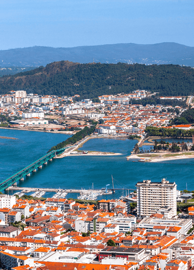 Aerial view of Viana do Castelo, Portugal, featuring a green bridge crossing the Lima River, a marina with docked boats, and a mix of orange-roofed buildings and modern apartments, with lush hills in the background.