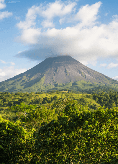 Arenal Volcano in Costa Rica with its peak partially covered by clouds, surrounded by lush green tropical forest under a blue sky.