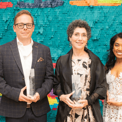 Group photo of award recipients and University of Guelph Alumni Association at the Art Gallery of Guelph. Seven people stand side by side, some holding awards and framed certificates, with a colourful mosaic background