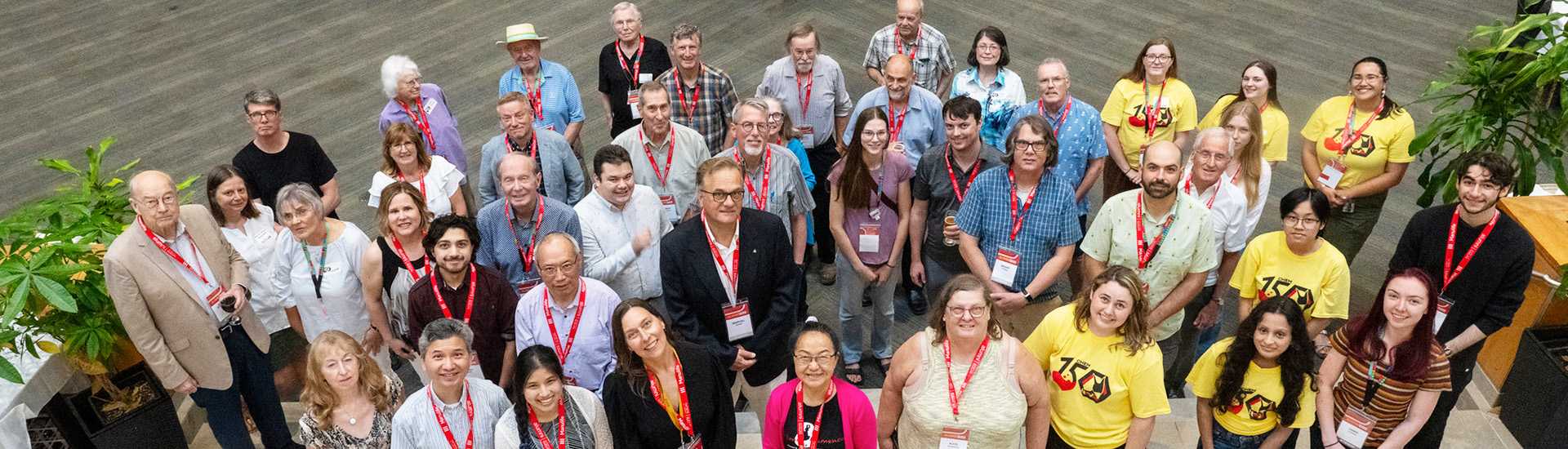 A diverse group of University of Guelph alumni and current students gather for a group photo during an alumni event. They are smiling and wearing red lanyards with name tags, while some are in yellow t-shirts with a celebratory logo. The group is standing together in a spacious indoor area with greenery in the background.