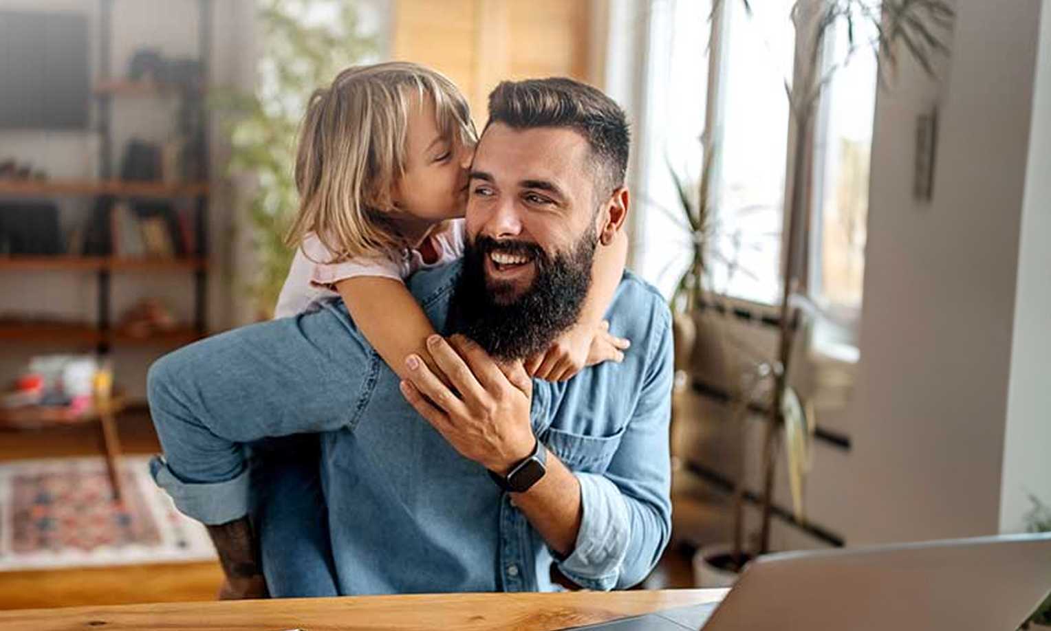 A young daughter kissing her bearded father on the cheek as he smiles, with a laptop visible on the desk in a cozy home office setting.