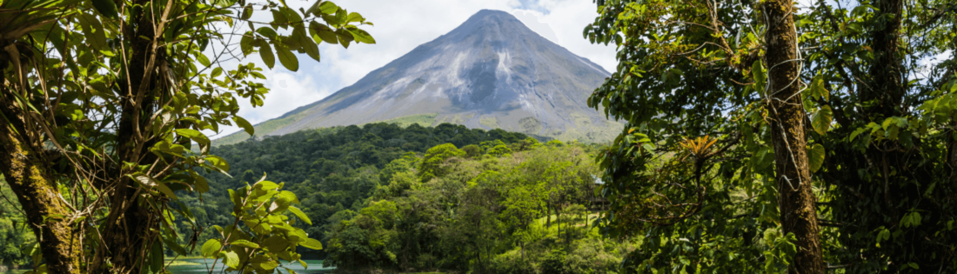 : A panoramic view of Arenal Volcano in Costa Rica, surrounded by lush tropical rainforest. The towering volcano rises above the dense greenery, framed by vibrant foliage under a partly cloudy sky.