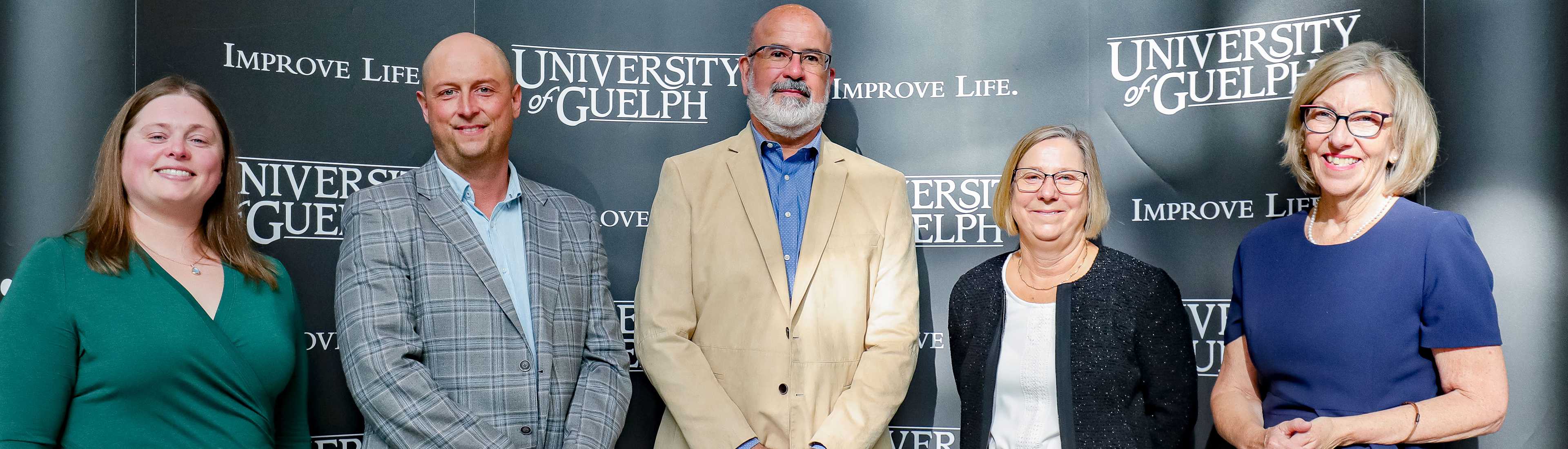 Group photo of four individuals standing in front of a University of Guelph backdrop with the slogan 'Improve Life.' The group includes three women and one man, all smiling and dressed in business attire