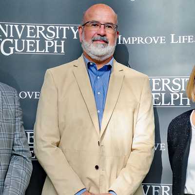 Group photo of four individuals standing in front of a University of Guelph backdrop with the slogan 'Improve Life.' The group includes three women and one man, all smiling and dressed in business attire