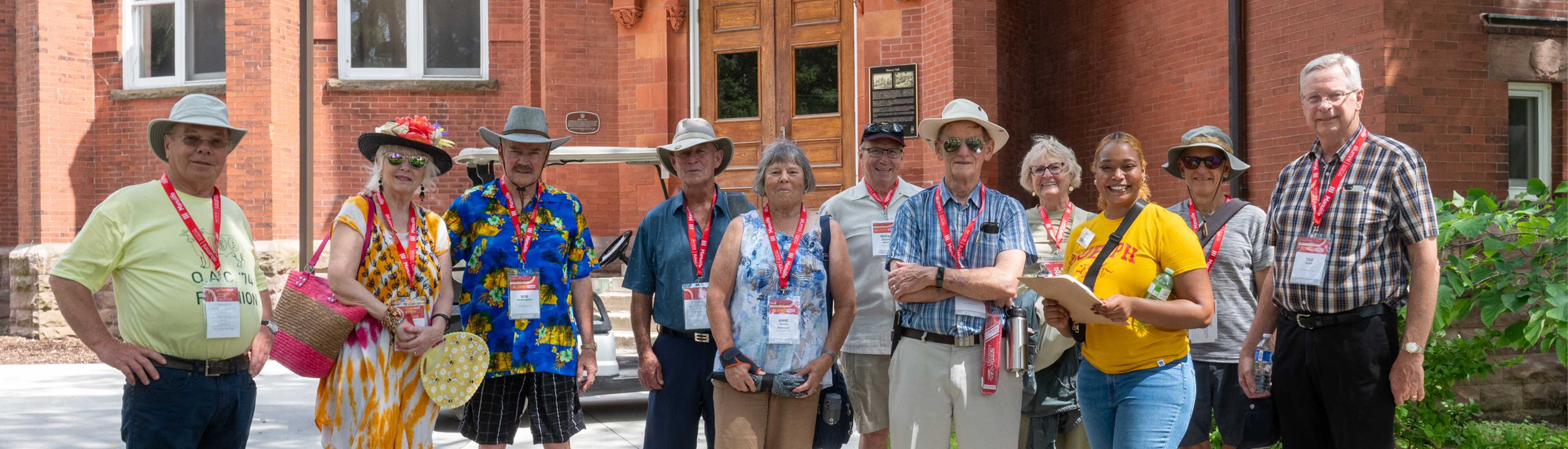 A group of older adults wearing name tags and red lanyards pose for a photo outside a historic red-brick building. They are dressed in colorful, casual summer attire, including sun hats, sunglasses, and floral prints. A young woman in a bright yellow "Guelph" T-shirt stands among them, holding a clipboard and smiling. The group appears to be on a guided campus tour, with some carrying water bottles and bags. The background features a wooden door, plaques on the wall, and green foliag